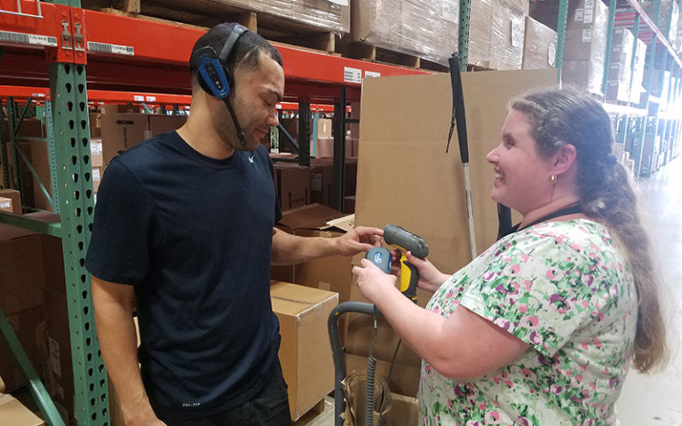 Two people work side by side in a warehouse, one wearing a headset while the other passes handheld equipment.