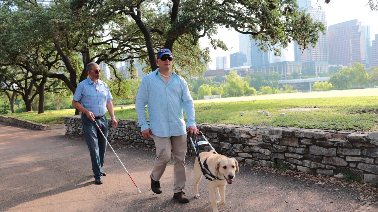 Two people walk along an outdoor path together, one using a white cane and the other with a guide dog