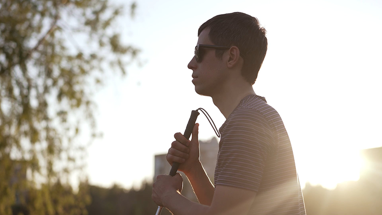 A person standing in profile against a bright outdoor background. They’re confident and calm, wearing a striped shirt and sunglasses, holding a white cane upright in front of them. The photo has a circular crop.